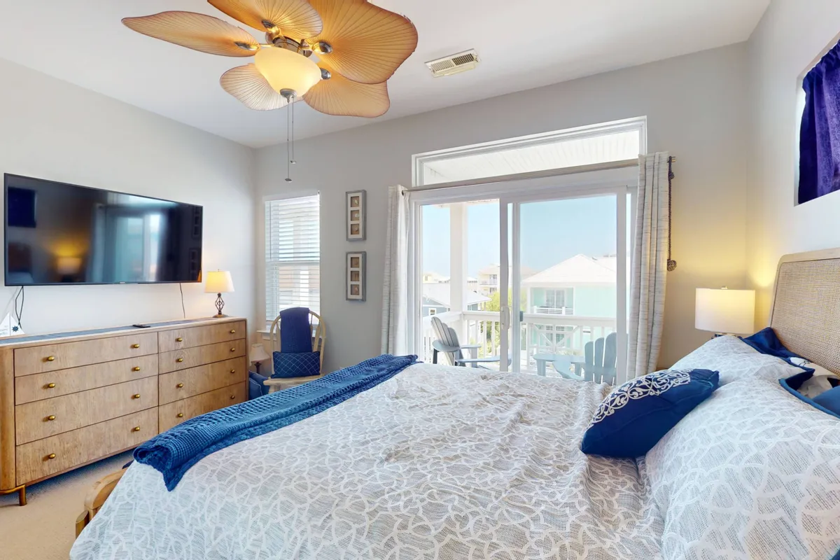 A spacious bedroom featuring a large bed neatly made with fresh linens and decorative pillows. The room includes a wall-mounted television, a ceiling fan, and a sliding glass door that opens to a private balcony with outdoor seating, offering views of the surrounding landscape. Natural light streams in, creating a tranquil atmosphere.