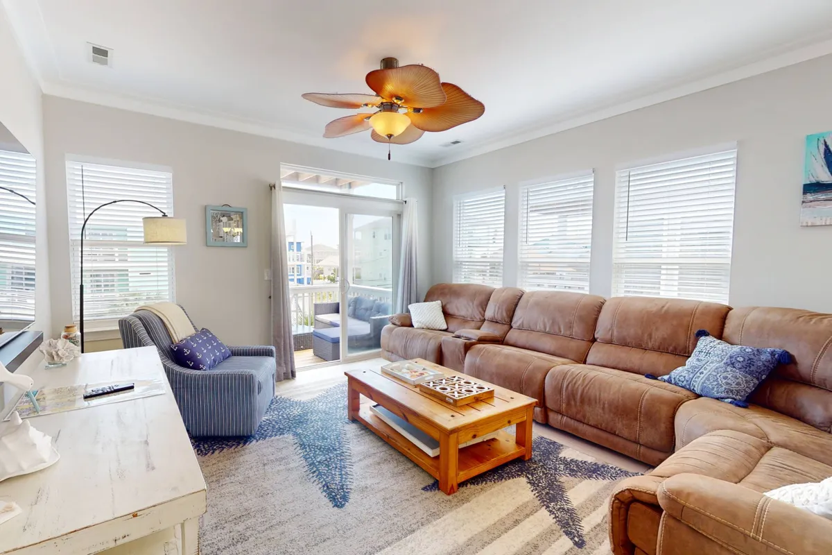 A spacious, sunlit living room with a large sectional sofa arranged around a coffee table, offering plenty of seating for gatherings. Decorative wall art adds a touch of character, and a large sliding glass door opens to an outdoor balcony, inviting natural light and ocean views into the space.