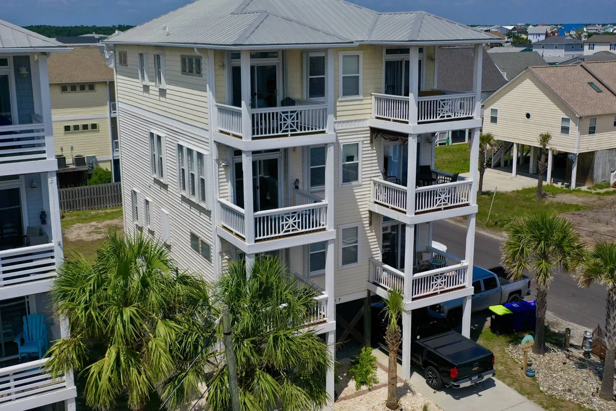 The image displays the exterior of a vacation home framed by swaying palm trees, featuring multiple balconies and exterior stairs. People are visible relaxing on balconies, with subtle hints of family gatherings and outdoor dining. The scene is lit with warm, natural light, suggesting the possibility of sunset views and a cheerful, vacation-like mood.