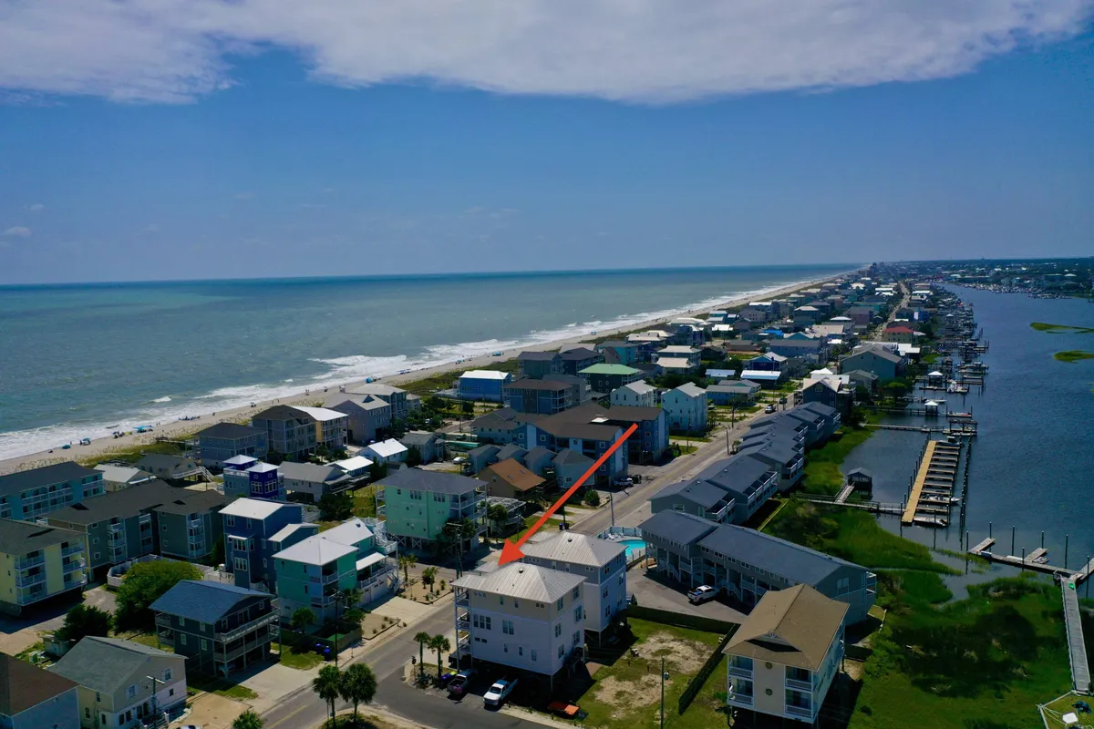 This image shows a beachfront property with the ocean in the background and neighboring houses nearby. The angle highlights both the proximity to the water and the tranquil, scenic surroundings, capturing the serene atmosphere at the north end of Pleasure Island. Soft lighting suggests either sunrise or sunset, enhancing the romantic mood.