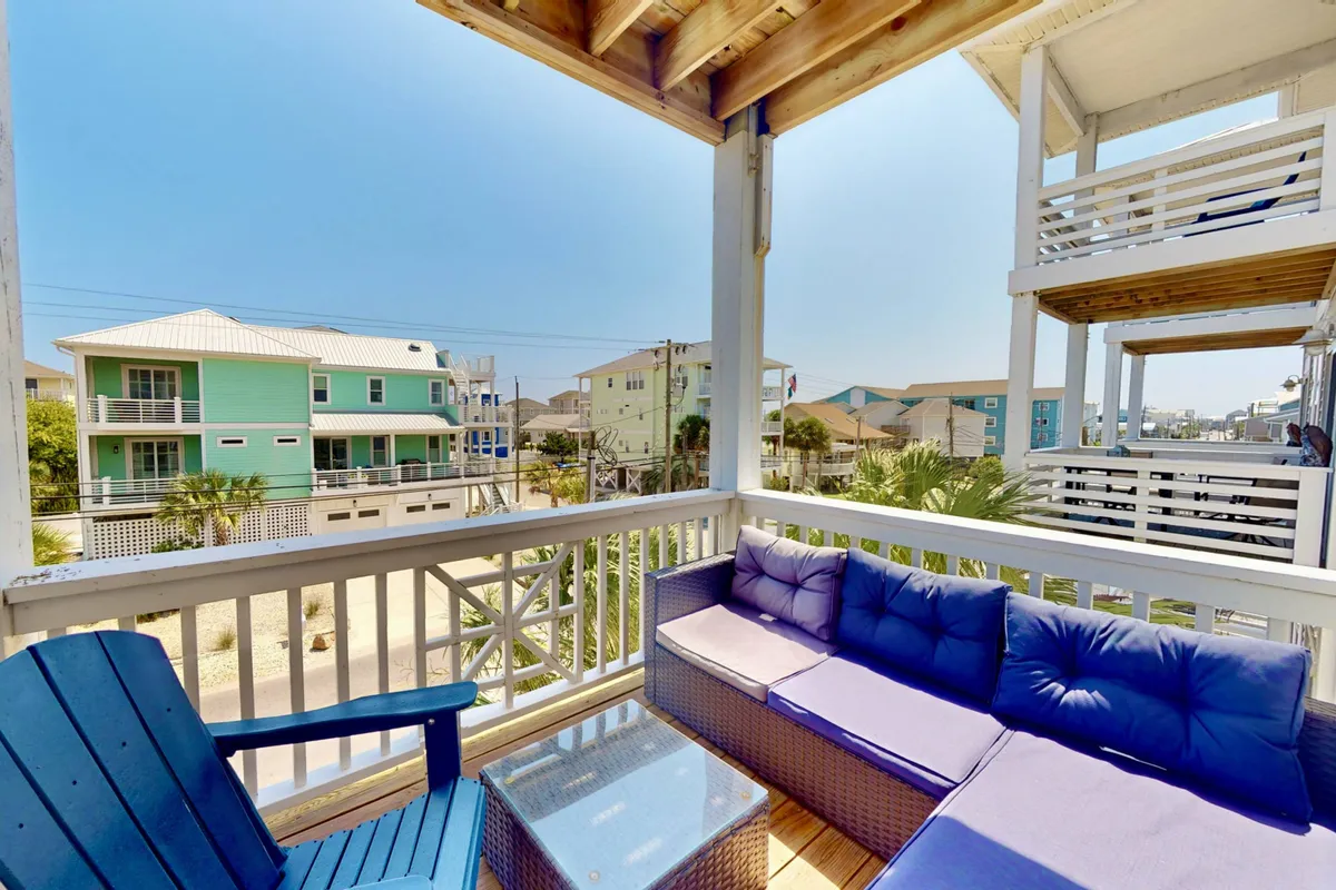 The hero image showcases the exterior of the Anchor Down townhouse, with inviting blue outdoor furniture set against a backdrop of palm trees and neighboring homes. The balcony is positioned perfectly for sunset views, and the outdoor space is designed for relaxing, enjoying coffee, reading, or socializing. The overall mood is tranquil and casual, capturing the laid-back coastal vibe of Carolina Beach.
