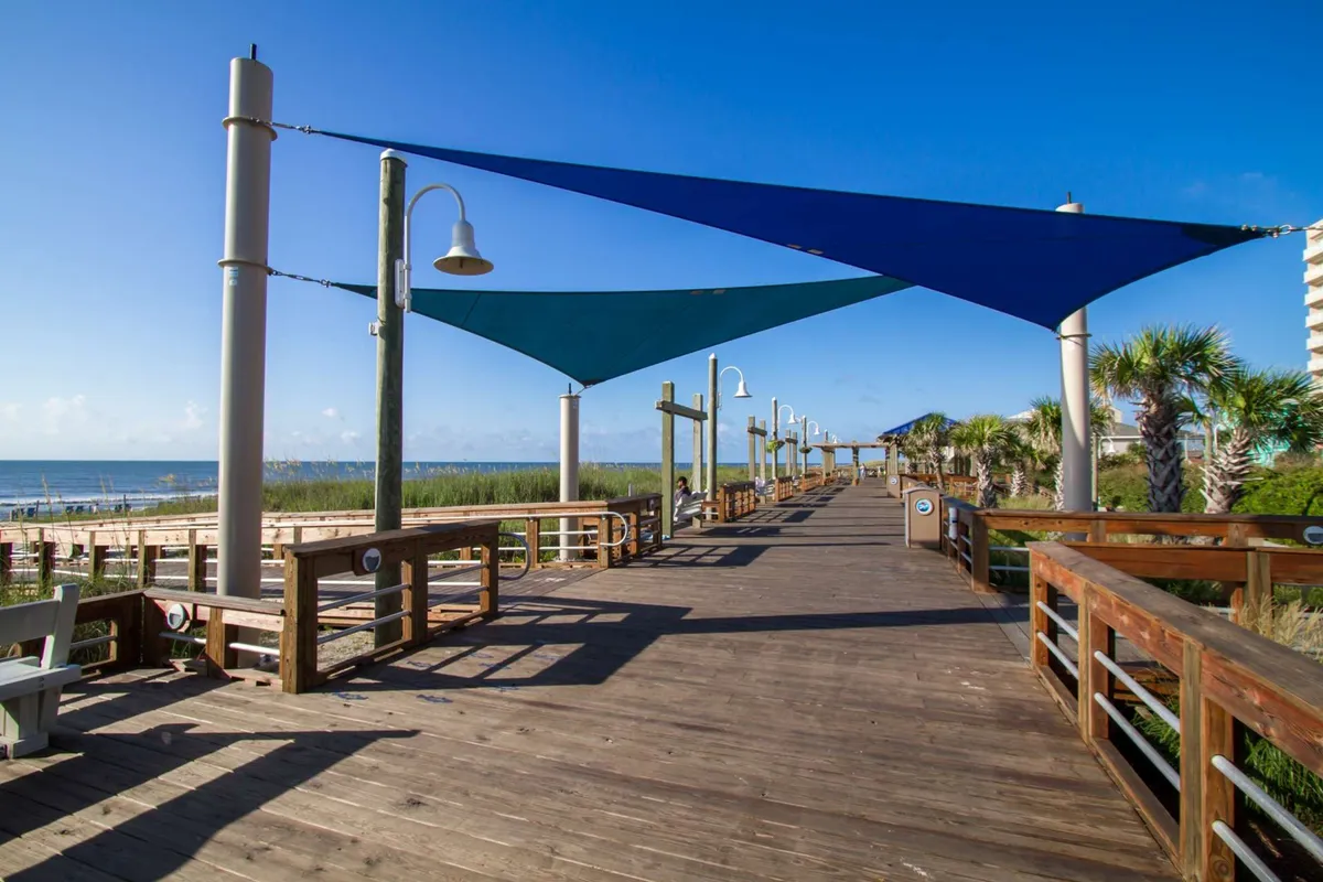 This photo showcases a wooden boardwalk adorned with bright shade sails and flanked by palm trees, leading directly to a beautiful beach view. Families and children can be imagined walking here, setting the scene for fun outings to the boardwalk’s attractions.