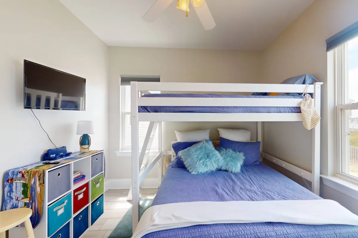 A cheerful bunk room with a colorful bunk bed, plenty of plush pillows, a vibrant storage unit, and a window letting in daylight. A television is positioned for entertainment, and the decor is playful, making the space inviting for children.
