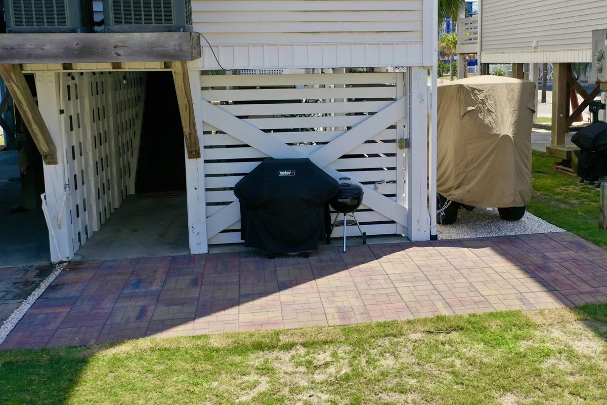 A paved patio area with a visible grill and relaxed outdoor seating is pictured. The scene suggests informal gatherings, barbecuing, and sharing meals outdoors. The area is partially shaded and feels private, aligning with the narrative of an exclusive ground-level patio for the group.