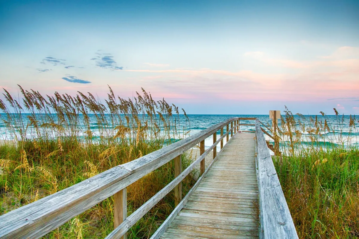 A serene exterior shot featuring a wooden boardwalk leading through beach grass toward the ocean with gentle waves and a colorful sky overhead. The setting sun casts a warm, romantic glow, inviting quiet evening strolls and peaceful reflection.