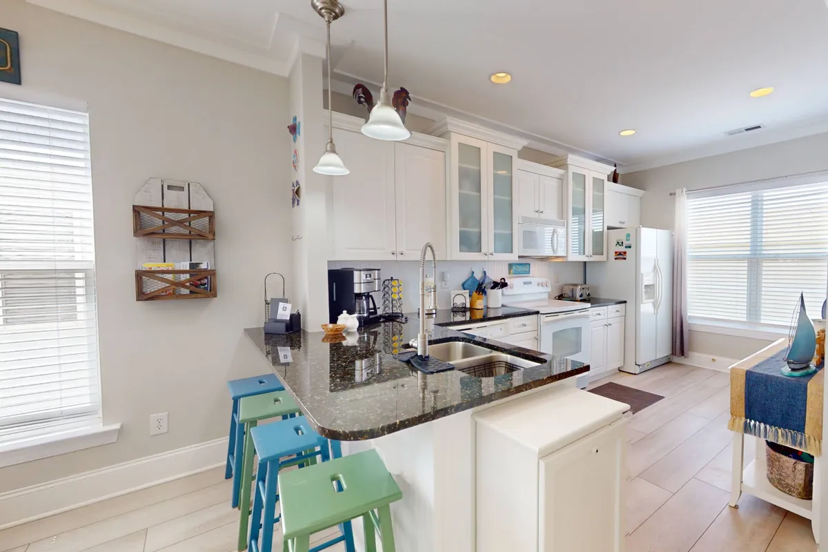 A modern kitchen featuring a granite countertop, colorful bar stools, and sleek appliances. Sunlight streams through windows, illuminating a welcoming space ideal for cooking and socializing, with ample room for group activity.