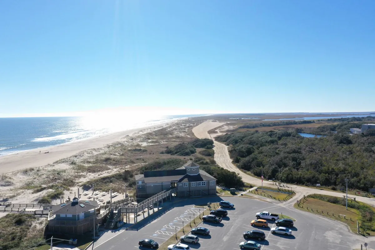 This image captures the beach, ocean, adjacent dunes, parking area, and the boardwalk, providing a sweeping view of the immediate surroundings and direct access points to the sand. People can be seen making their way to the shore, and the landscape feels welcoming and ready for exploration.