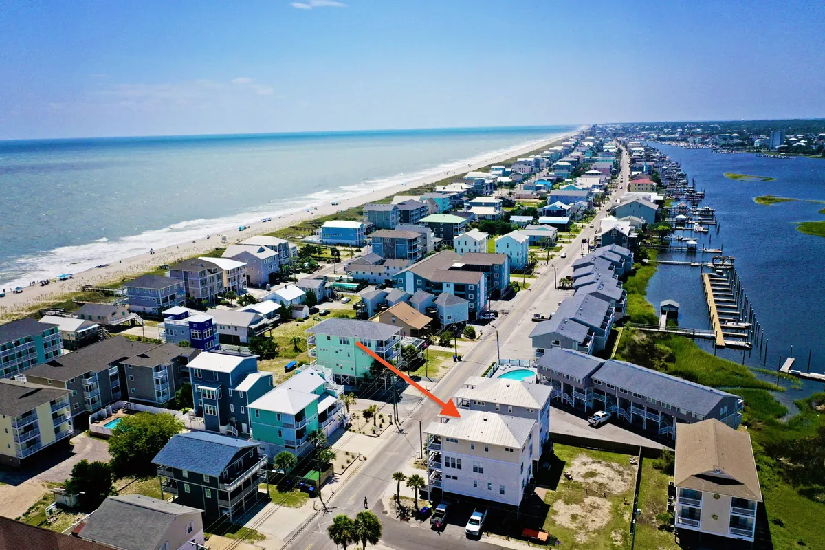 A wide view of the beachfront area with colorful houses, boats in the marina, and a relaxed beach scene in the foreground. The composition includes both water and vibrant local architecture, suggesting proximity to both the beach and area attractions like restaurants and shops.