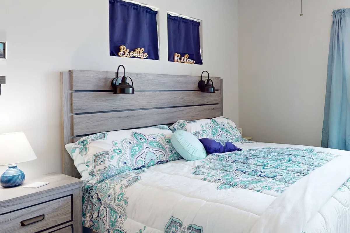 A tranquil queen bedroom with a neatly made bed featuring decorative bedding and pillows, soft lighting from a bedside lamp, and uplifting wall decor. The ambiance is peaceful and inviting, ideal for restful nights.