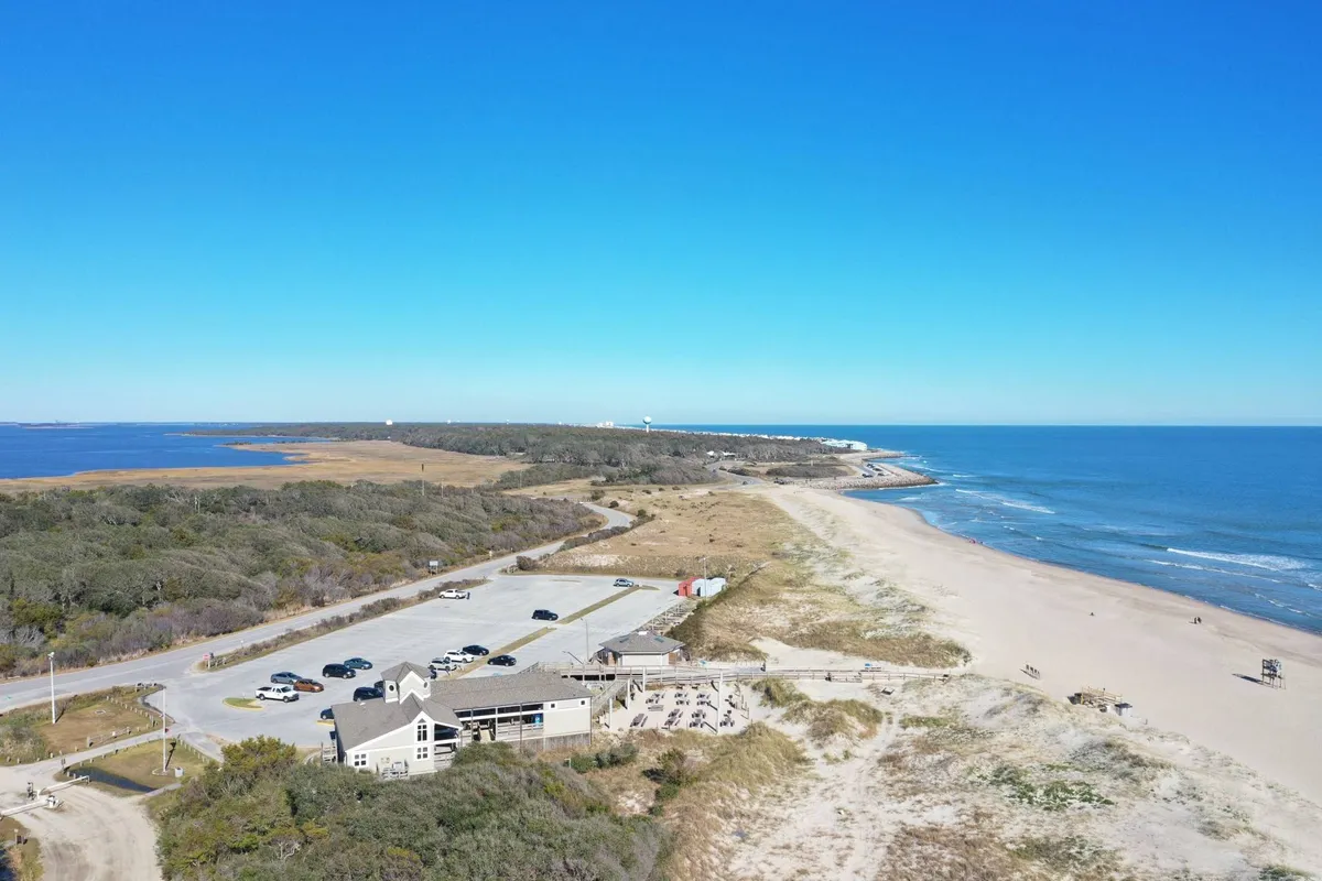A wide view of a sandy beach stretching alongside the ocean, bordered by a parking area, nearby vegetation, and a coastal structure. The scene highlights an accessible and inviting shoreline, perfect for outdoor activities such as sunbathing, swimming, fishing, hiking, and wildlife observation.